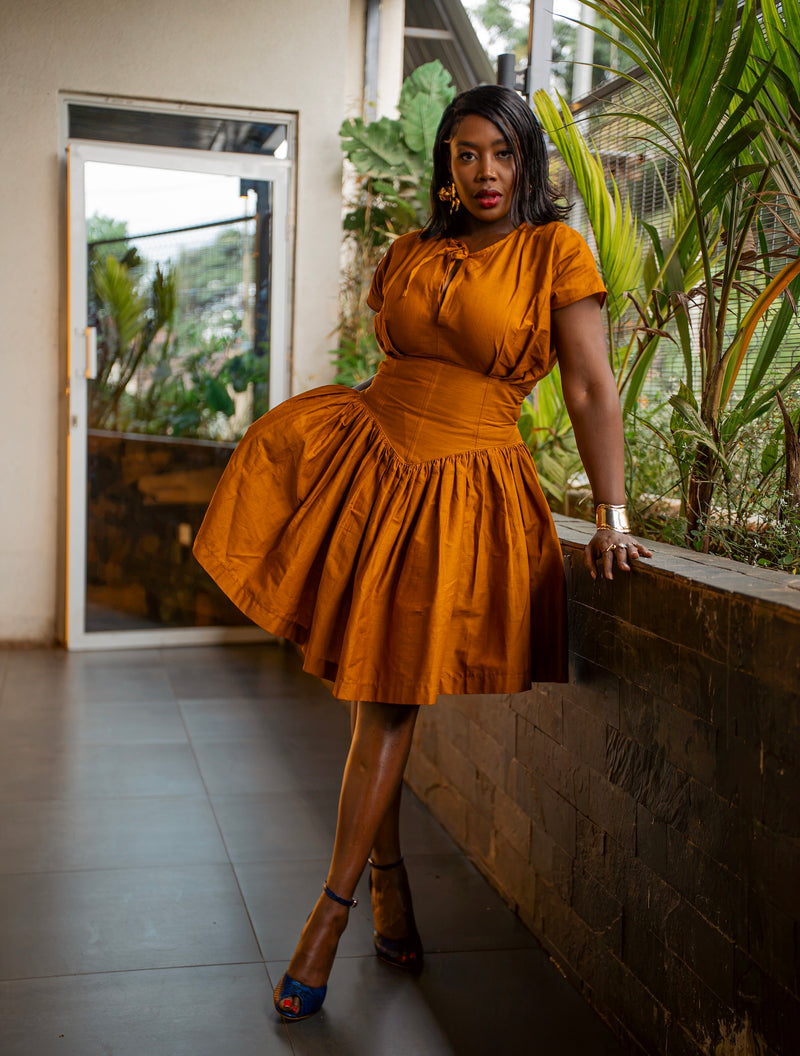 Mustard UMBO short dress with a tie at the neckline, structured corset-style waistband and gathered skirt, leaning on a stone planter wall